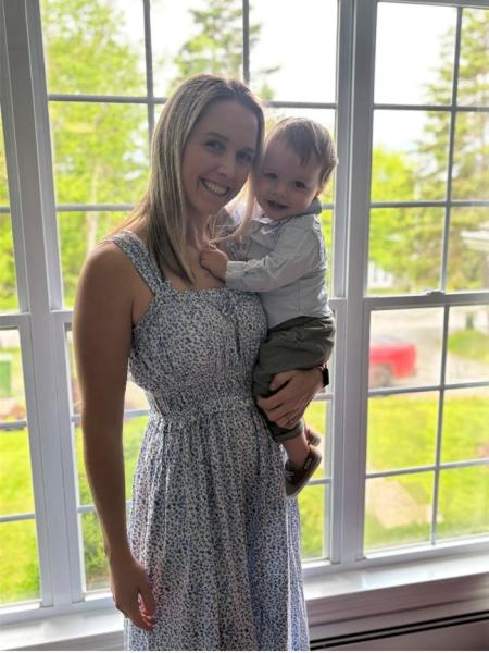 A woman with blonde hair stands in front of a window wearing a white dress with a floral print, holding a baby, and is smiling at the camera.