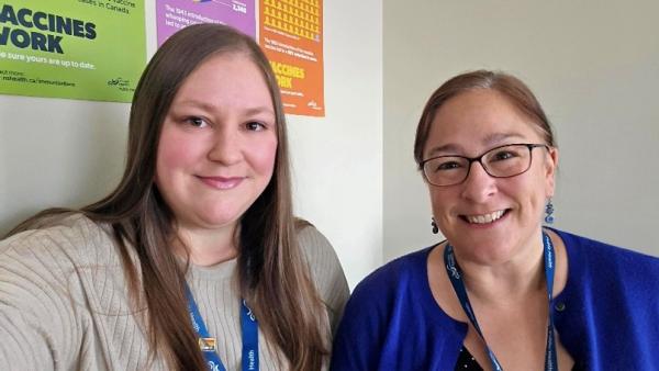 Two women sit side-by-side, one has long blonde hair and the other has brown hair in a ponytail and glasses.