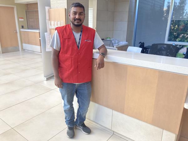 A man with a red best and jeans stands at a front desk in the lobby of a healthcare centre, smiling at the camera.