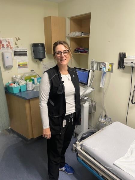 A person wearing glasses, a white long-sleeve shirt, and a black vest is standing in a medical examination room beside medical equipment and supplies.