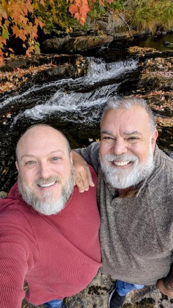 Two men standing in front of a waterfall with a fall leaves. Man on the left has a gray beard, wearing a red sweater. Man on the right has gray hair and gray beard, wearing a brown/grey sweater. 