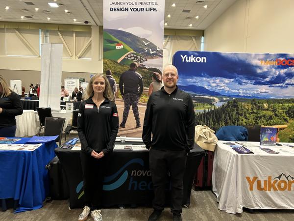 Woman standing on the left, man on the right. Both wearing black and they are standing in front of information tables.