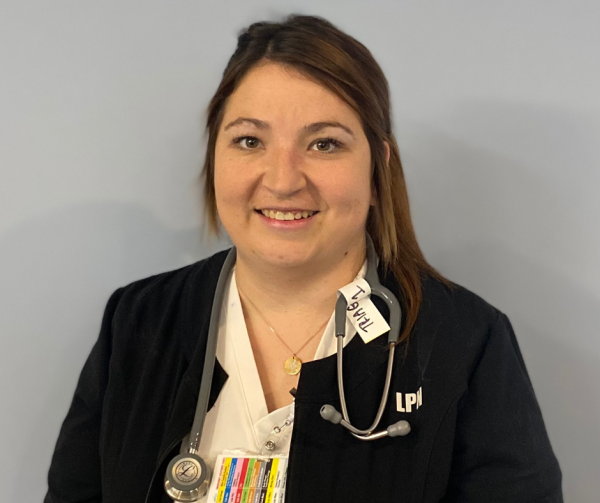 A healthcare worker wearing a black jacket with “LPN” on it smiles at the camera. She has a stethoscope around her neck and stands in front of a plain light-coloured background.  