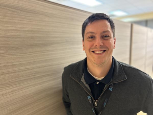 A man with short brown hair stands against a wood-panel wall wearing a zip-up sweater and a golf shirt, and is smiling at the camera.