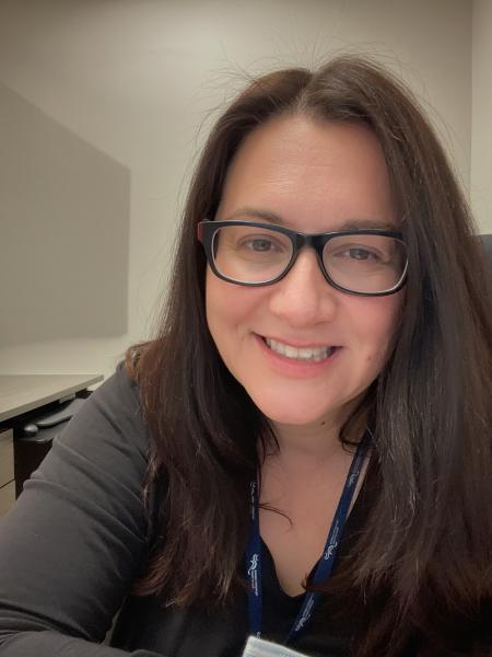 A woman with glasses and brown hair wears a brown shirt and is smiling at the camera.