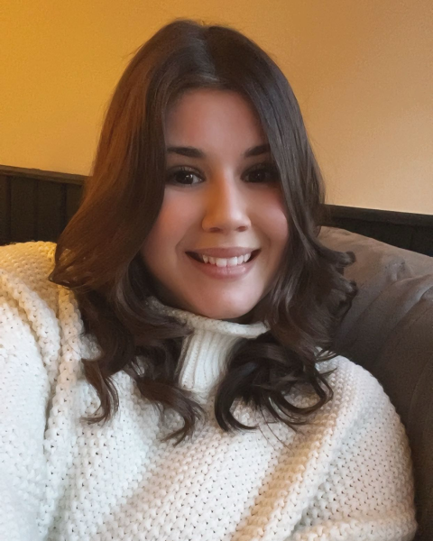 A woman with brown hair wears a white sweater and is sitting on a chair, smiling at the camera.