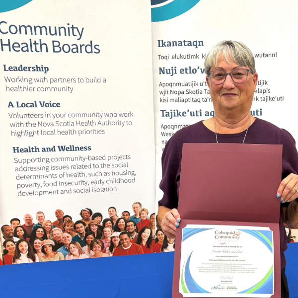 In an indoor setting, a smiling woman holds a certificate standing in front of banners related to community health boards. 