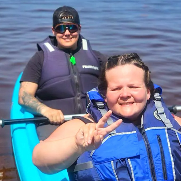 Two people in a kayak on a body of water. One person is sitting at the front, smiling and making a peace sign with their hand, while the other is seated behind them, wearing sunglasses and looking at the camera. Both are wearing life jackets. 