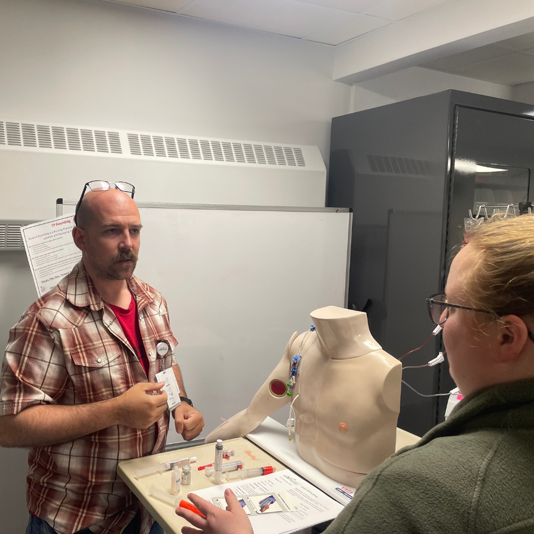 A man with a plaid shirt wearing glasses on his head is standing in front of a woman and is showing her a simulated medical procedure.