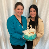 Two people stand together smiling and holding a cake that is blue with two swans on top. 