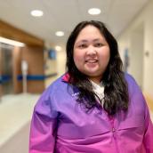 A person with long brown hair is standing facing the camera, they are wearing a pink and blue jacket, smiling at the camera.
