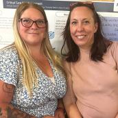 Two women are sitting and smiling at the camera. One on the left has long blonde hair and glasses with a flower printed shirt, and the other has medium-length brown hair and a peach shirt on.