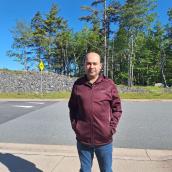 A photo of a man with short black hair. He is wearing a maroon sweater and jeans. He is standing outside with trees in the background. 