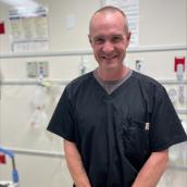 Man stands in a hospital room wearing a black scrub shirt and is smiling at the camera.