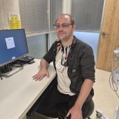 A man sits at a desk wearing a black zip-up sweater, a stethoscope, a beige shirt, and glasses and is smiling at the camera.