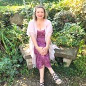 A woman sitting on a bench outside wearing a purple flower pattern dress, highlighting her smile and warm expression. 