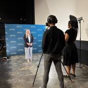 A woman stands in front of a blue backdrop with a camera person, camera and lighting person standing in front of her.