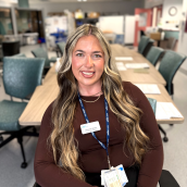 A woman with long wavy blonde and brown hair sits smiling in a meeting room. She is wearing a dark long-sleeve top, a name badge, and a blue lanyard with ID cards. The background shows a long conference table, green chairs, and hospital equipment in a bright clinical setting.