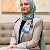 A photo of a woman wearing a teal blue hijab with a beige pant suit and a black shirt underneath. She has her name badge on her collar and a green stethoscope. 