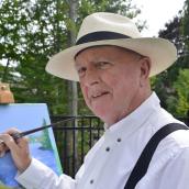 Man wears a white, button-up shirt, suspenders, and a tan and black hat while painting outdoors with trees behind him.