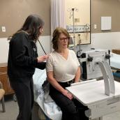 A healthcare provider assists a seated woman on a hospital examination table using a stethoscope in a clinical room.