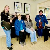 Four women pose for a photo, gather around a piano. One person holds a guitar; another person holds fiddle. The other two women sit on the piano bench. 