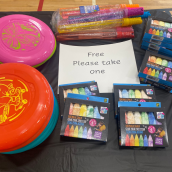 A sign reading “free please take one” displays on a table, indoors, filled with various colourful objects. 
