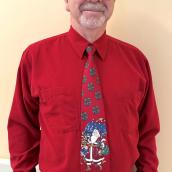 In an indoor setting, a smiling man wearing glasses is standing facing the camera. Also, he is wearing a red shirt and tie. The tie has an image of Santa Claus on it. 