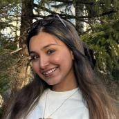 Surrounded by trees, a woman smiles at the camera in an outdoor setting.
