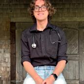 A smiling man wearing glasses, along with casual clothing - such as denim jeans and a top with sleeves - sits on an outdoor bench. A building is visible in the background. 