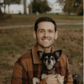Photo of a man outside with his dog. He is wearing a brown plaid jacket and has brown hair. The dog is black and brown with a brown collar.