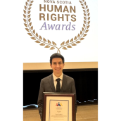 A man, smiling, dressed in a suit stands indoors holding a plaque. 