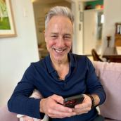 Photo of Robert Moffat siting on a couch at his residence and smiling at the camera while using the YourHealthNS app on his phone.