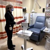 A masked healthcare worker holding a red clipboard stands beside an iPad on a workstation in a hospital exam room.