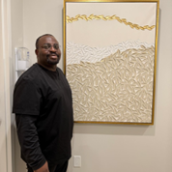 Dr. Adebayo stands in a clinic hallway beside a framed abstract artwork in neutral tones with gold accents, wearing black clinical attire and smiling toward the camera.