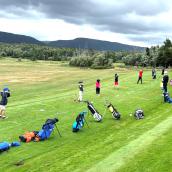 A group of people gathered on a golf course. The scene features lush grasslands under a clear sky with clouds and trees in the background. 