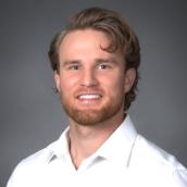 A portrait setting of a smiling man indoors wearing a white shirt with a plain wall in the background. 