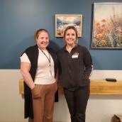 Two women stand together in a room painted blue with framed photos of flowers on the wall. They are wearing casual work wear and smiling at the camera.