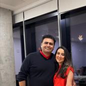 A photo of a man and a woman. The man is on the left and has brown hair. He is wearing a black sweater over a red collared shirt. The woman is on the right and she has long brown hair. She is wearing a red blouse with black pants.