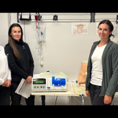 Five healthcare staff stand in a clinical training room beside peritoneal dialysis equipment and a simulation mannequin. Tubing, IV lines, and dialysis supplies are set up on a table and wall behind them, showing hands-on dialysis education in a hospital setting.