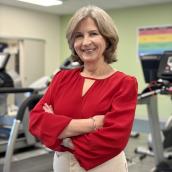 A woman stands in a gym wearing a red shirt and beige pants with her arms folded, smiling at the camera.