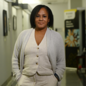 A woman stands in a hallway wearing a white vest, pants and grey sweater, smiling at the camera.