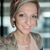A woman with short, blonde hair wears a pink shirt with a printed sweater over top and is smiling at the camera.