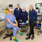 A group of four medical professionals sit in a hospital room wearing scrubs and surrounded by medical equipment.