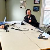 Three people wearing headsets are seated indoors in a room at a table with microphones. 