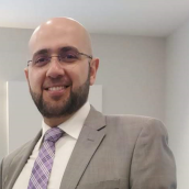  A man wearing a light gray suit jacket, a white dress shirt, and a purple patterned tie stands indoors against a plain, light‑colored wall with minimal décor.