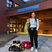 Image of a nurse in black and white scrubs, wearing a stethoscope around her neck and a firefighter jacket over her right shoulder. She is facing the camera smiling. She has fire fighter pants and boots, plus a medical bag next to her feet. She is standing outside a hospital "Eastern Shore Memorial Hospital."