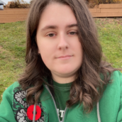 A photo of a woman outside with brown wavy hair. She is wearing a green zip-up sweater with a grey design on the left shoulder and a red poppy pin.