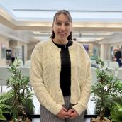 A woman stands in a room wearing a white cardigan, black shirt and grey pants, smiling at the camera.