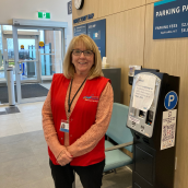 A woman stands in the lobby of a healthcare facility wearing a red vest, pink shirt, black pants, glasses and a smile.